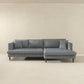 The Ashcroft Furniture Blake Velvet Upholstered L-Shape Chaise Sofa Sectional 107 in modern gray stands against a plain light beige wall and floor, adding comfort and style to a minimal, spacious room.