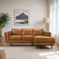 A modern living room features the Ashcroft Furniture Chase MCM Tan Leather Chaise Sofa Sectional 92, paired with a large potted plant, floor lamp, and a framed mountain landscape above the sofa, bathed in natural light from the window.