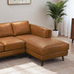 The Ashcroft Furniture Chase MCM Tan Leather Chaise Sofa Sectional 92 with wooden legs sits on a beige rug in a bright mid-century modern living room, with a potted green plant and a wooden cabinet in the background.