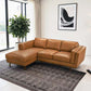 A modern living room with the Ashcroft Furniture Chase MCM Tan Leather Chaise Sofa Sectional 92, a patterned dark gray rug, a large potted plant near tall windows, and abstract black-and-white art on the wall.