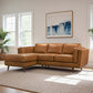 A modern living room featuring the Ashcroft Furniture Chase MCM Tan Leather Chaise Sofa Sectional 92, a large beige rug, two sunlit windows, a potted plant, and an abstract painting on the wall.