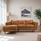 A modern living room features the Chase MCM Tan Leather Chaise Sofa Sectional 92 by Ashcroft Furniture, a floor lamp, a large potted plant, and a framed mountain landscape photo. Sunlight streams through sheer white curtains at the window.