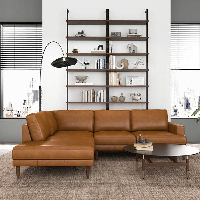 A modern living room with the Ashcroft Furniture Corbin MCM Tan Leather Sectional Chaise Sofa 108, a mid-century round dark wood coffee table, floor lamp, tall open bookshelf with decor, and large windows showcasing a cityscape view.