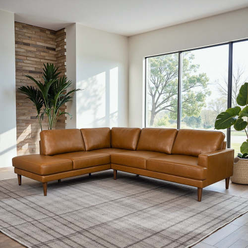 A modern living room features the Ashcroft Furniture Corbin MCM Tan Leather Sectional Chaise Sofa 108 on a gray plaid rug, with large windows, potted plants, and a mid-century modern stone accent wall in the corner.