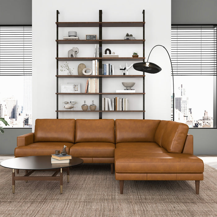 A modern living room features the Ashcroft Furniture Corbin MCM Tan Leather Sectional Chaise Sofa 108, a round coffee table, tall black floor lamp, wall-mounted shelves with books and decor, and large windows showcasing a cityscape.