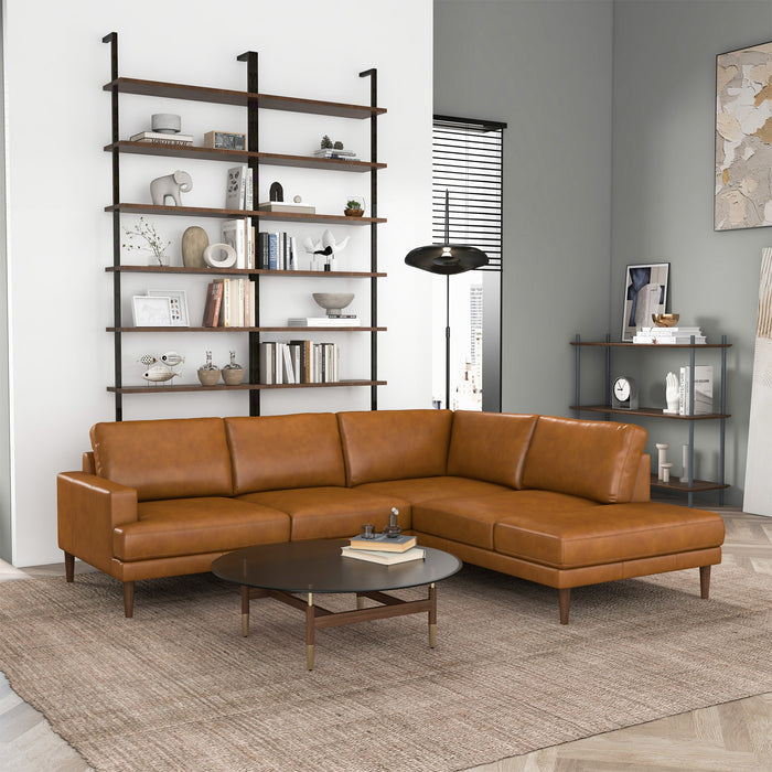 A modern living room featuring the Ashcroft Furniture Corbin MCM Tan Leather Sectional Chaise Sofa 108, a round coffee table, light brown rug, wall-mounted shelves, side table, and large windows for a mid-century modern vibe.
