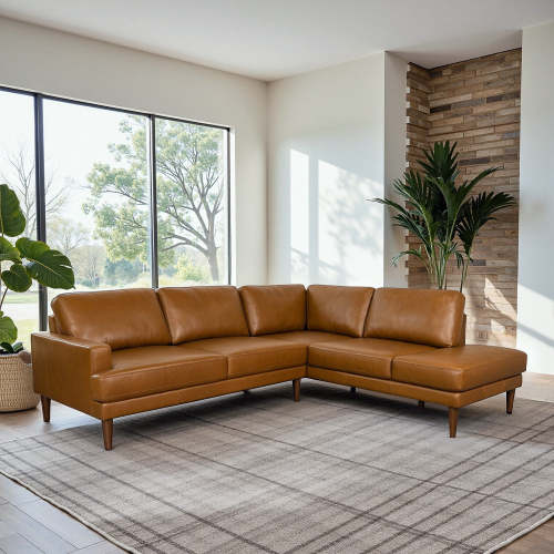 A modern living room featuring the Ashcroft Furniture Corbin MCM Tan Leather Sectional Chaise Sofa 108 on a gray patterned rug, with large windows, indoor plants, and a brick accent wall creates a cozy mid-century modern vibe.