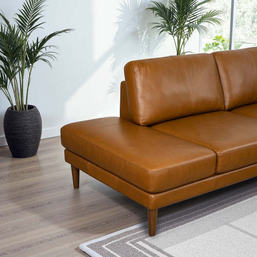 The Corbin MCM Tan Leather Sectional Chaise Sofa 108 by Ashcroft Furniture features a mid-century modern design with wooden legs, set on a light wooden floor near a window, complemented by potted green plants and a partially visible rug.