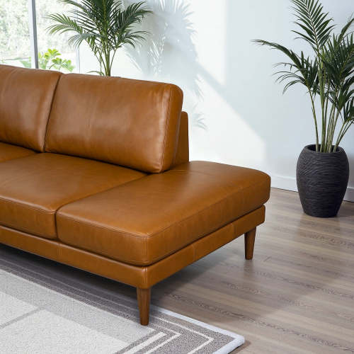 The Ashcroft Furniture Corbin MCM Tan Leather Sectional Chaise Sofa 108 with wooden legs sits on a patterned rug in a bright room. Green potted plants and sunlight streaming through the window complete the scene.