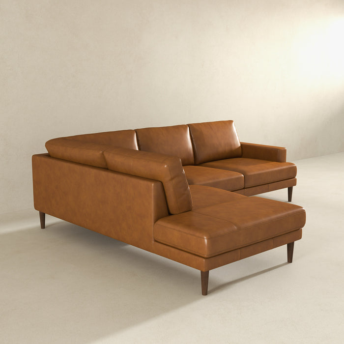 The Ashcroft Furniture Corbin MCM Tan Leather Sectional Chaise Sofa 108 features three seat and back cushions, a right-side chaise lounge, and wooden legs, perfectly complementing a minimalist room with light beige walls and flooring.