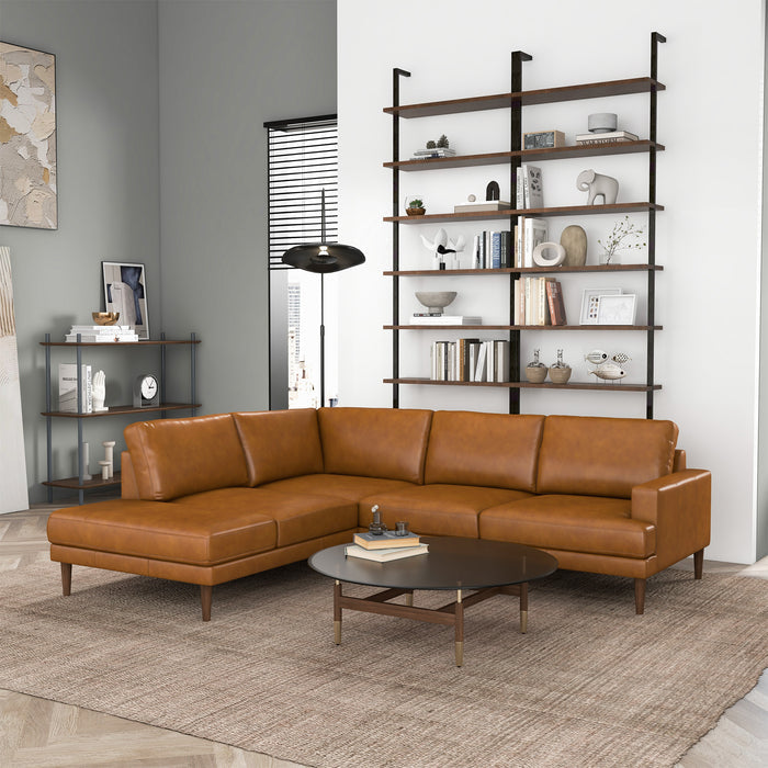 A modern living room featuring the Ashcroft Furniture Corbin MCM Tan Leather Sectional Chaise Sofa 108, a round coffee table, gray rug, wall-mounted shelves with books and decor, plus a console table with framed photos and a lamp.