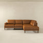 The Ashcroft Furniture Corbin MCM Tan Leather Sectional Chaise Sofa 108, with clean lines and wooden legs, sits against a plain off-white wall in a minimalistic room.