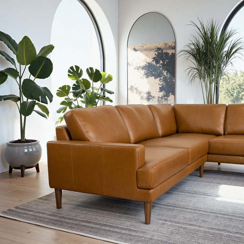 A modern living room showcases the Corbin MCM Tan Leather Sectional Chaise Sofa 108 by Ashcroft Furniture on a gray striped rug, with large green potted plants, arched windows, and an arched mirror reflecting a tree outside.